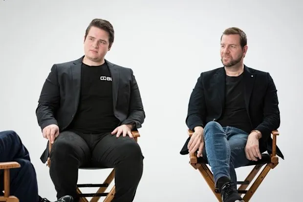 Two men seated in director's chairs against a white backdrop